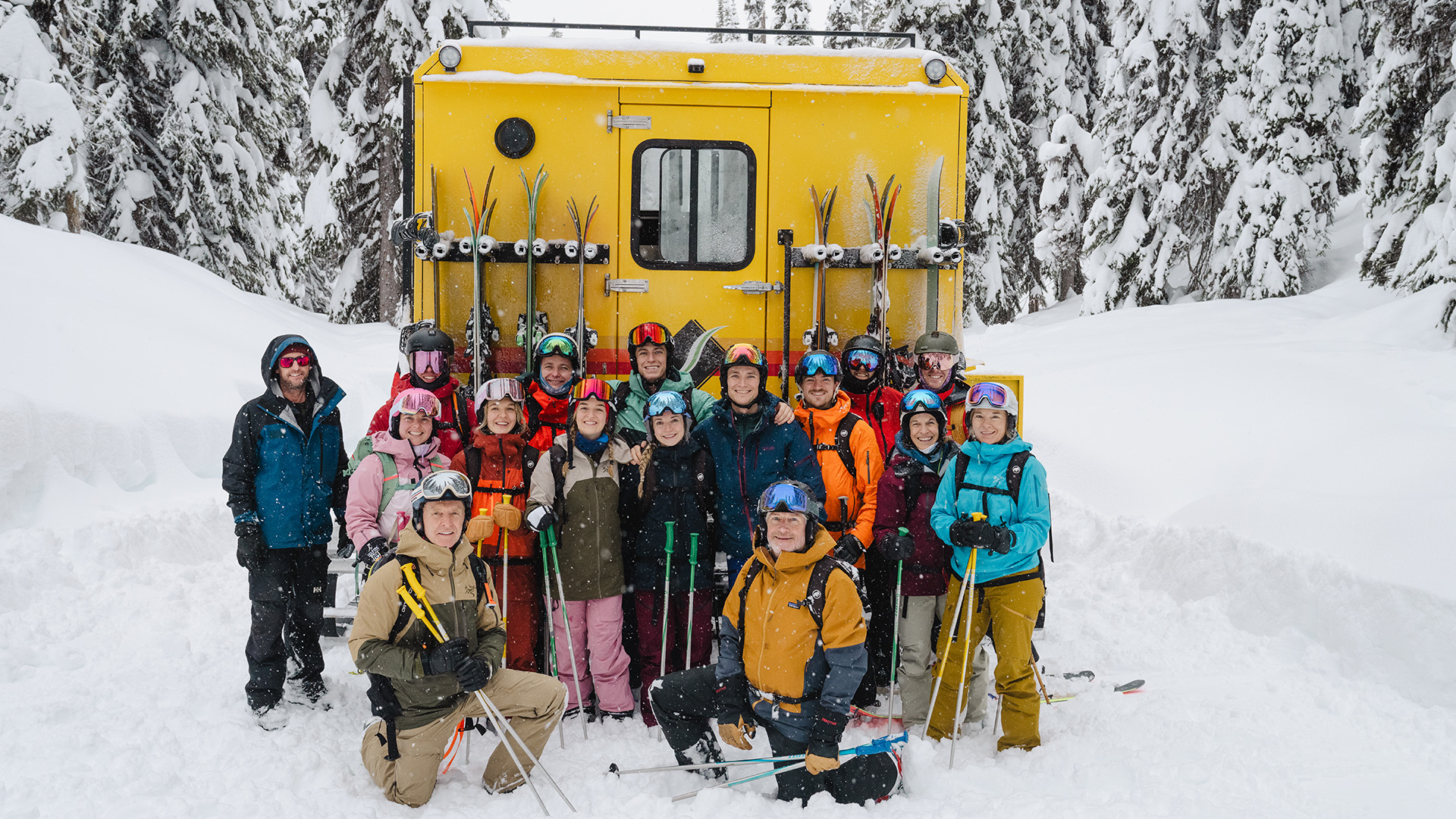 Group of skiers in colourful gear posing in front of a yellow snowcat during a guided backcountry ski trip.