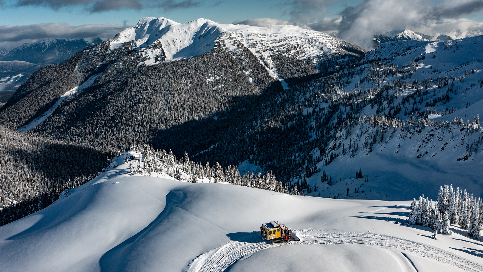 Yellow snowcat driving across untouched snow in the Canadian Rockies backcountry with mountain peaks in the distance.